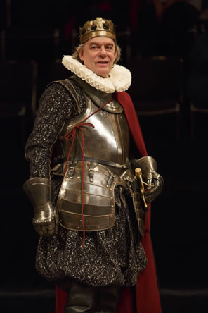King John in breast plate and hip armor, armored gloves over a black floral cloak, a ruff collar, a red cape, a sword at his hilt, and a simple gold crown